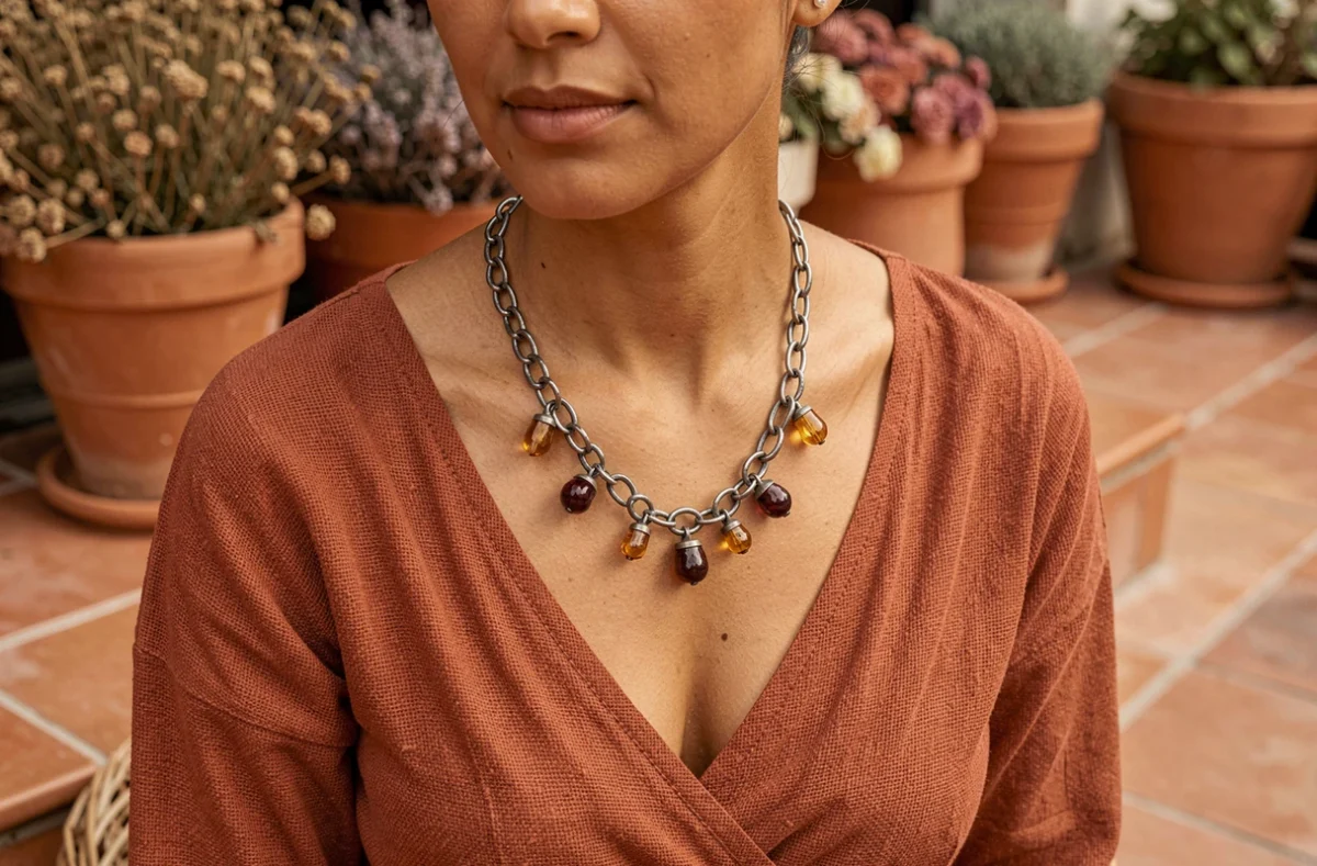 Femme à peau mate portant un collier bohémien en argent oxydé et gunmetal avec pendentifs ambre et bordeaux, haut en coton rouille, fond terrasse méditerranéenne